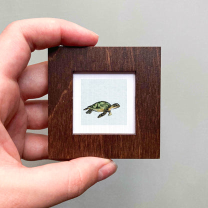 A hand is holding a small wooden frame containing a picture of a green turtle.