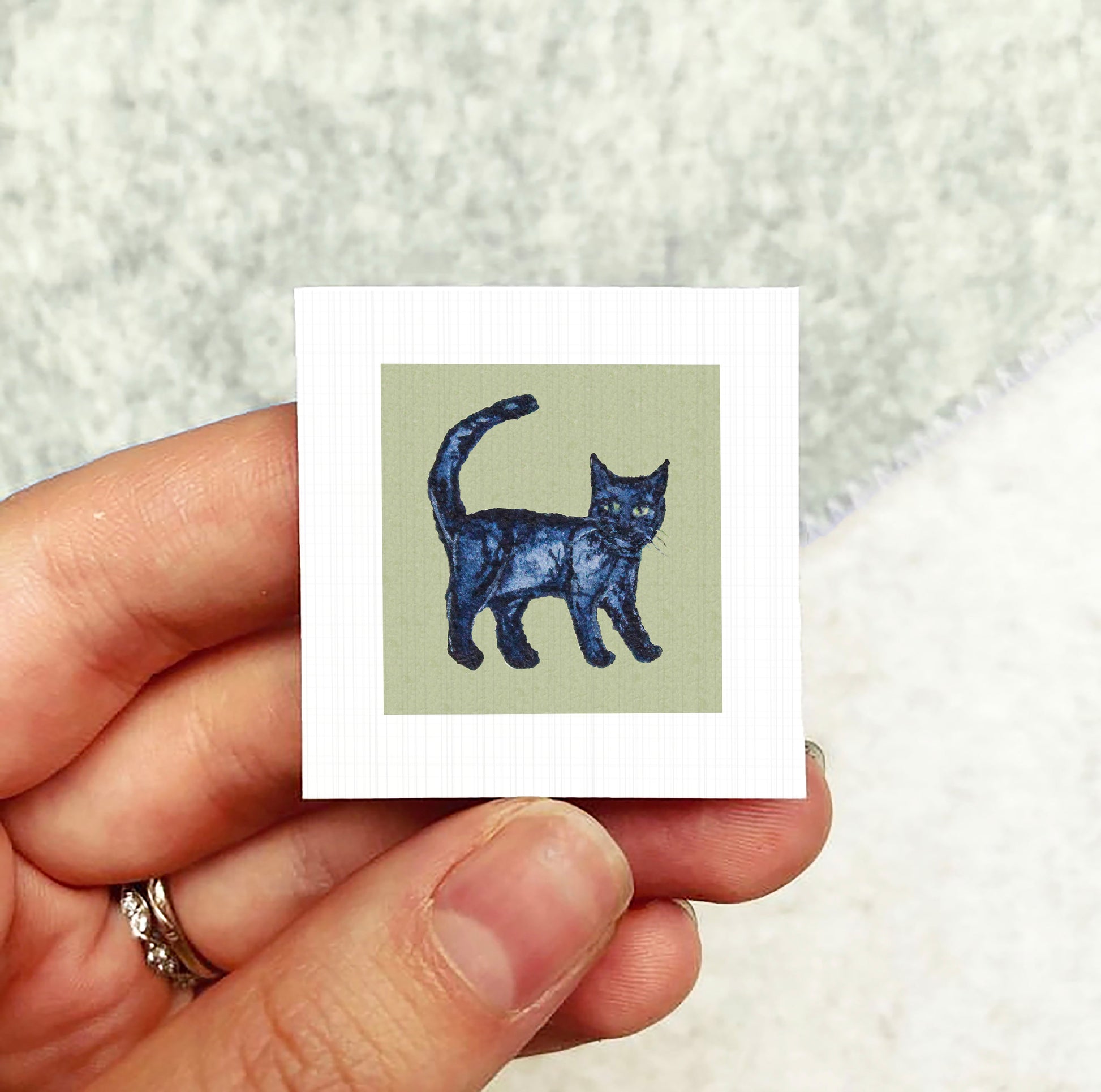 A hand holding a small square card with an image of a cat on it.