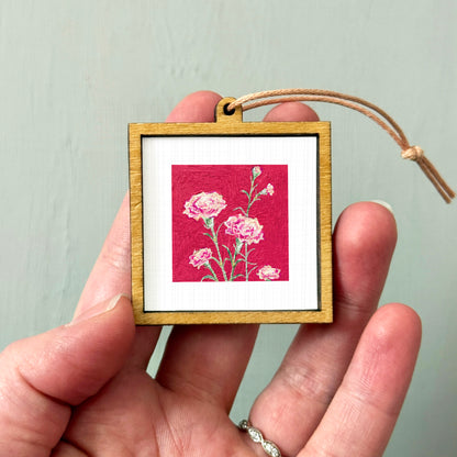 A hand holding a small wooden frame containing a painting of pink flowers against a red background.