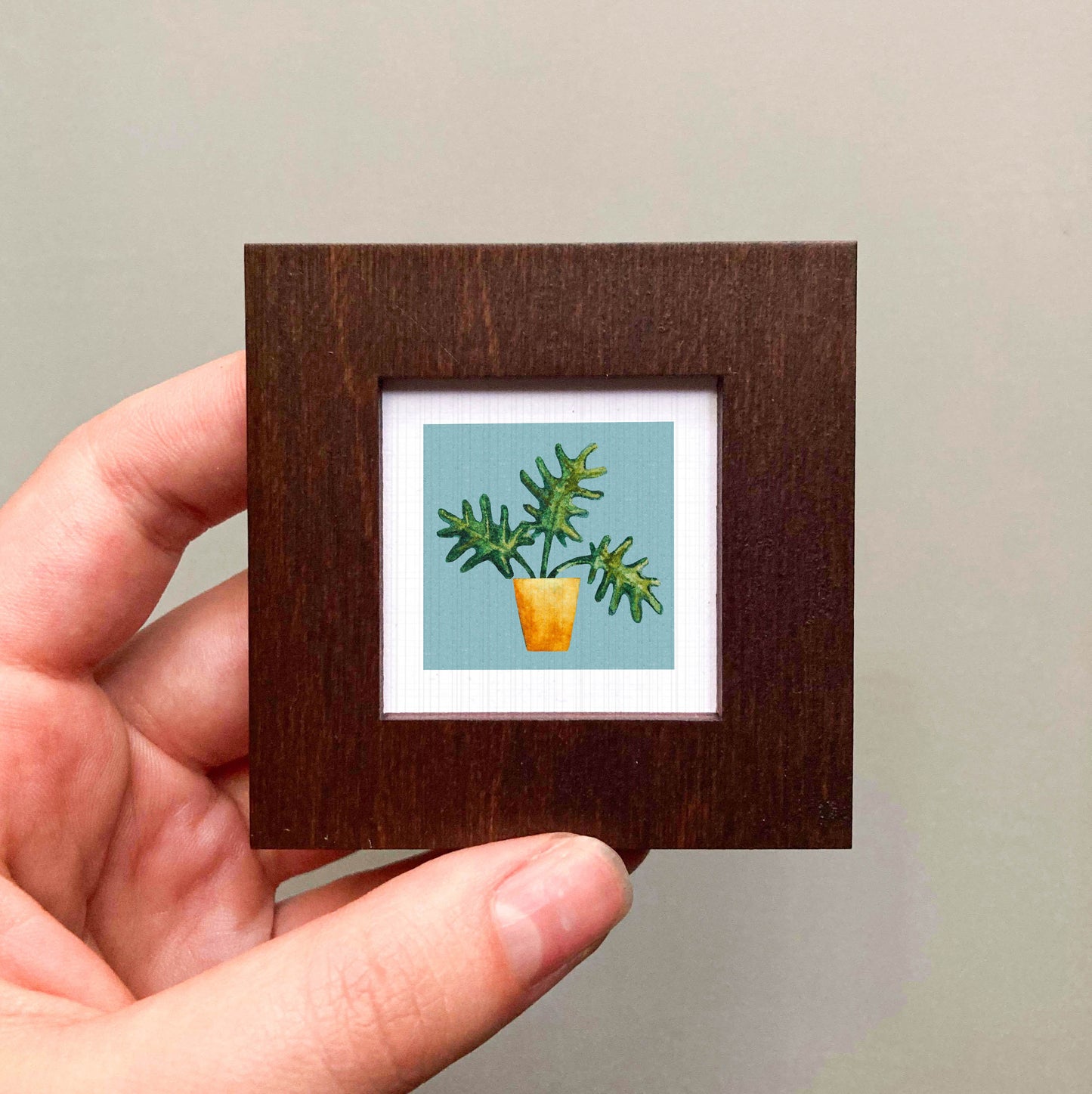 A hand is holding a small square frame containing a painting of a potted plant with green leaves.