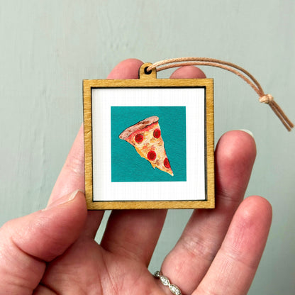 A hand holding a small wooden frame containing a picture of a slice of pizza.