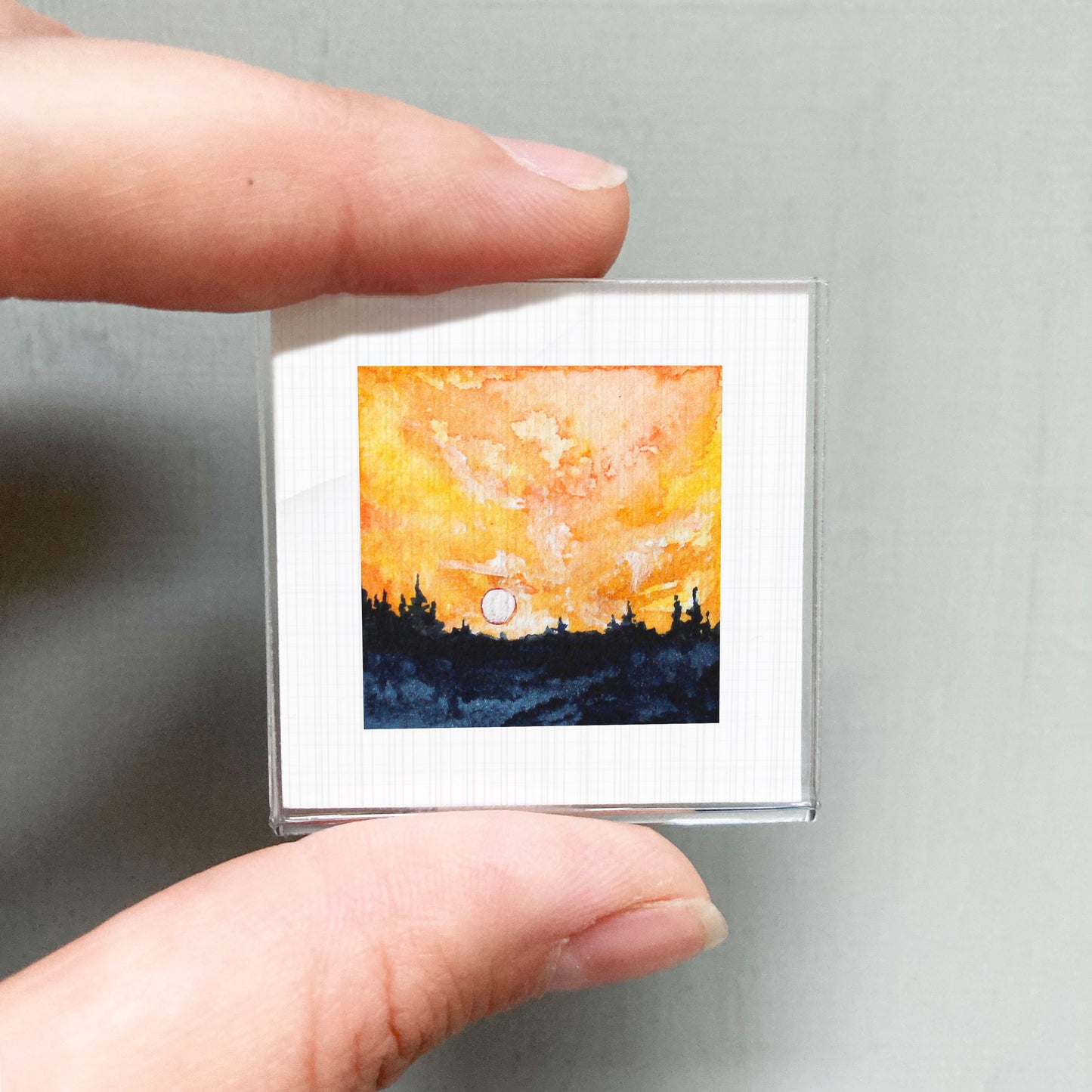 A person's hand is holding a small square picture depicting a sunset over a forest.