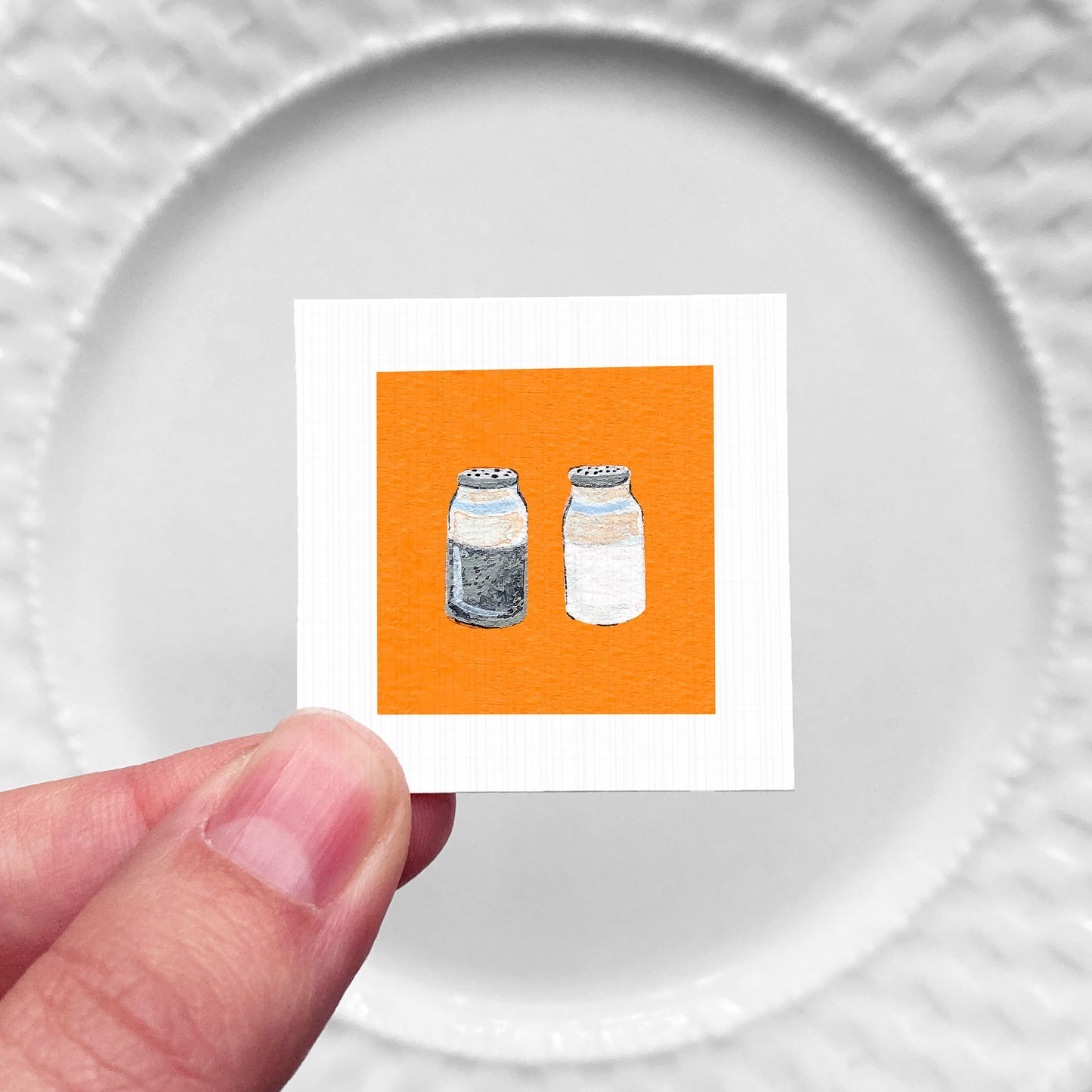 A hand is holding a small square with an illustration of two bottles, one black and one white, against a white background.