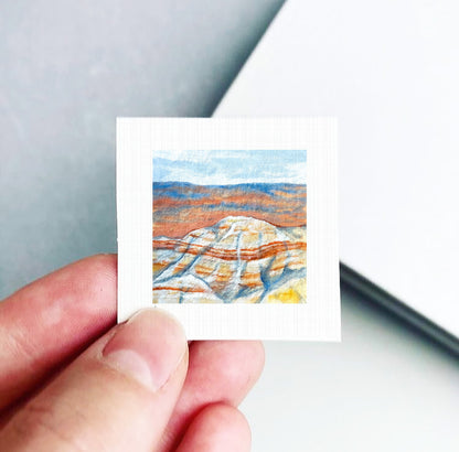 A hand is holding a small square piece of paper with a painting of a mountain landscape on it.
