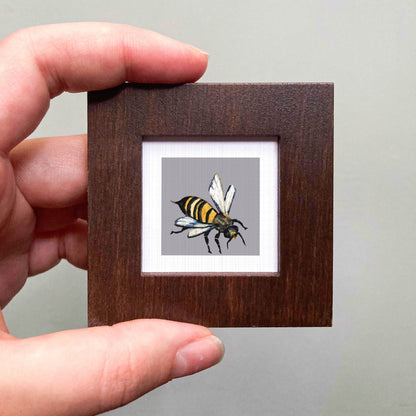 A person's hand is holding a small wooden frame containing a detailed illustration of a bee.
