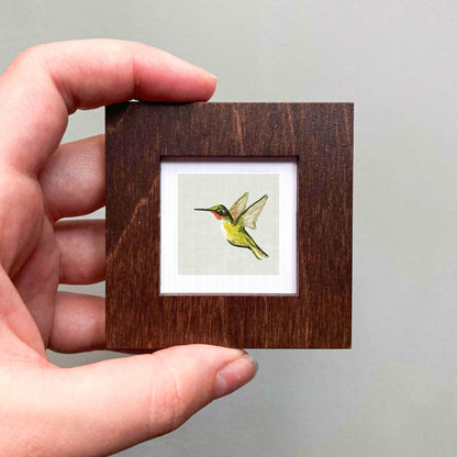 A hand is holding a small wooden frame containing a framed image of a hummingbird.