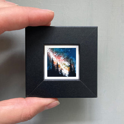 A hand is holding a small black frame containing a framed painting of a night sky with trees.