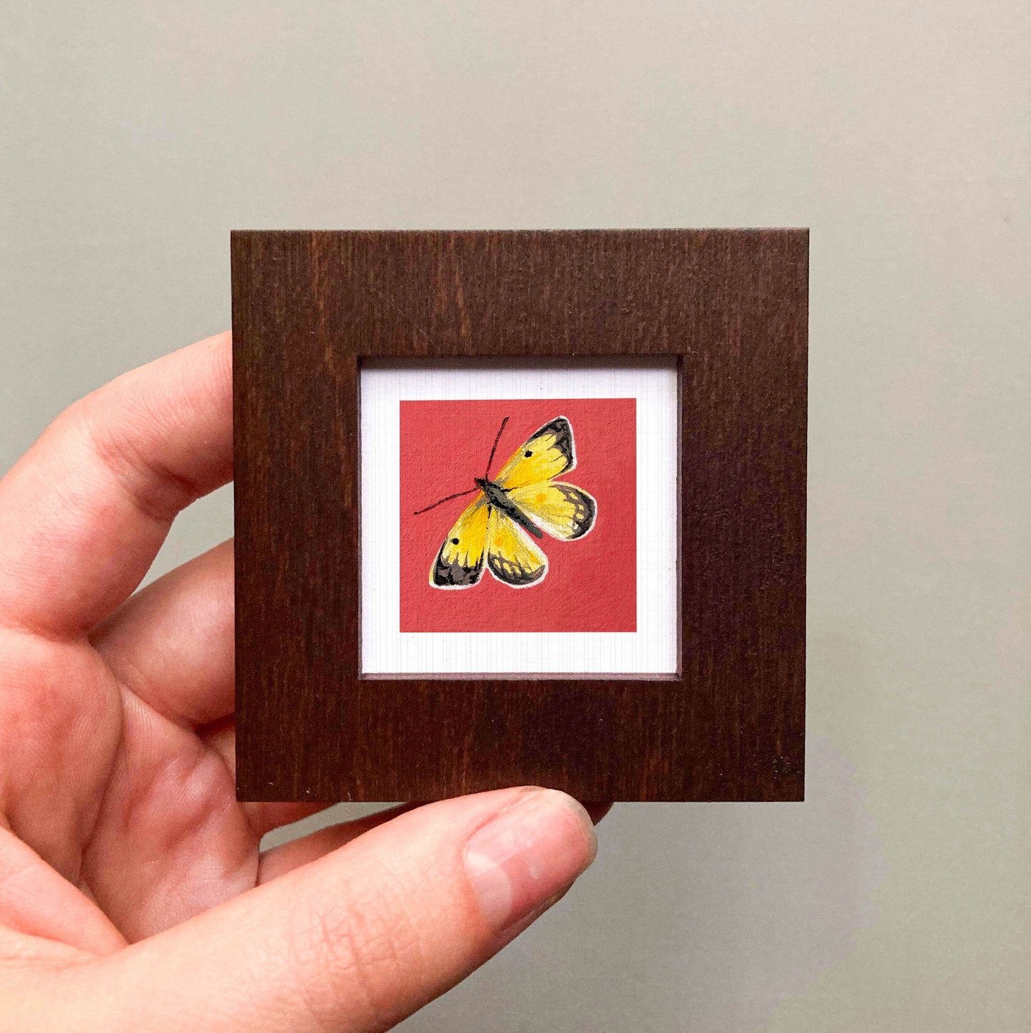 A hand is holding a small wooden frame containing a vibrant painting of a yellow butterfly with black markings.