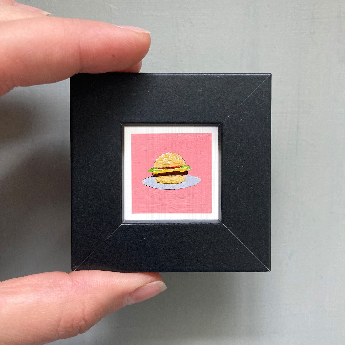 A person's hand is holding a small black frame containing a framed illustration of a hamburger.