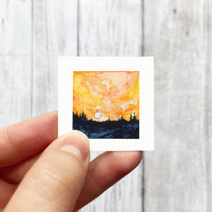 A hand is holding a small square painting depicting a sunset over a forest landscape.