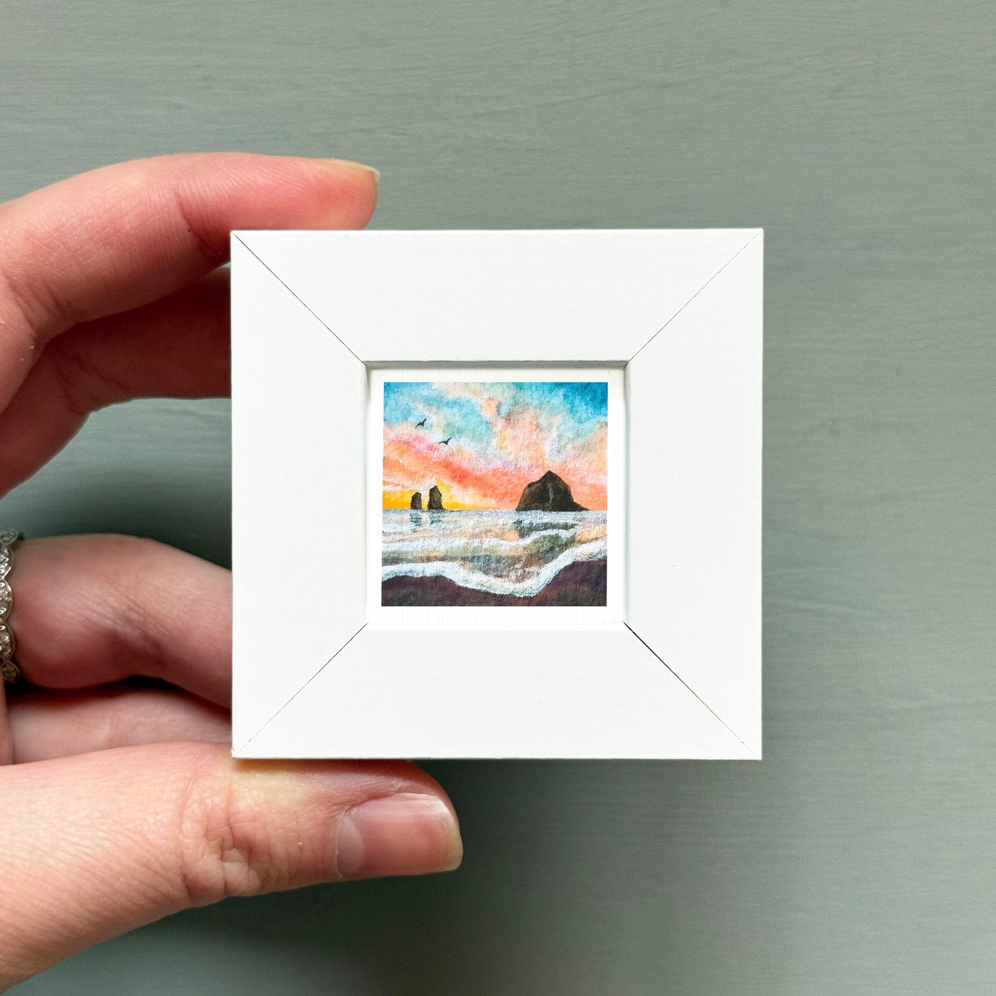 A hand is holding a small white square frame containing a painting of a landscape with a mountain and a body of water.
