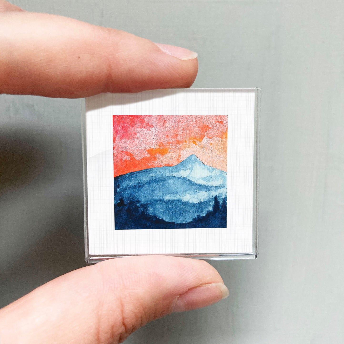 A hand is holding a small square picture of a mountain landscape with a vibrant sky.
