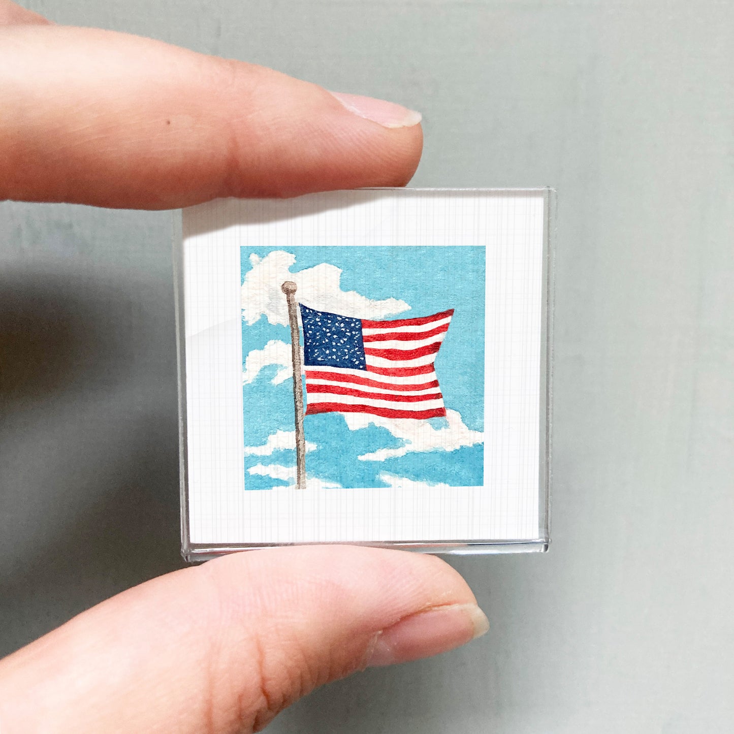 A person's hand is holding a small square-shaped object that displays an American flag against a cloudy sky background.