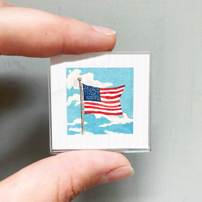A person's hand is holding a small square-shaped object that displays an American flag against a cloudy sky background.