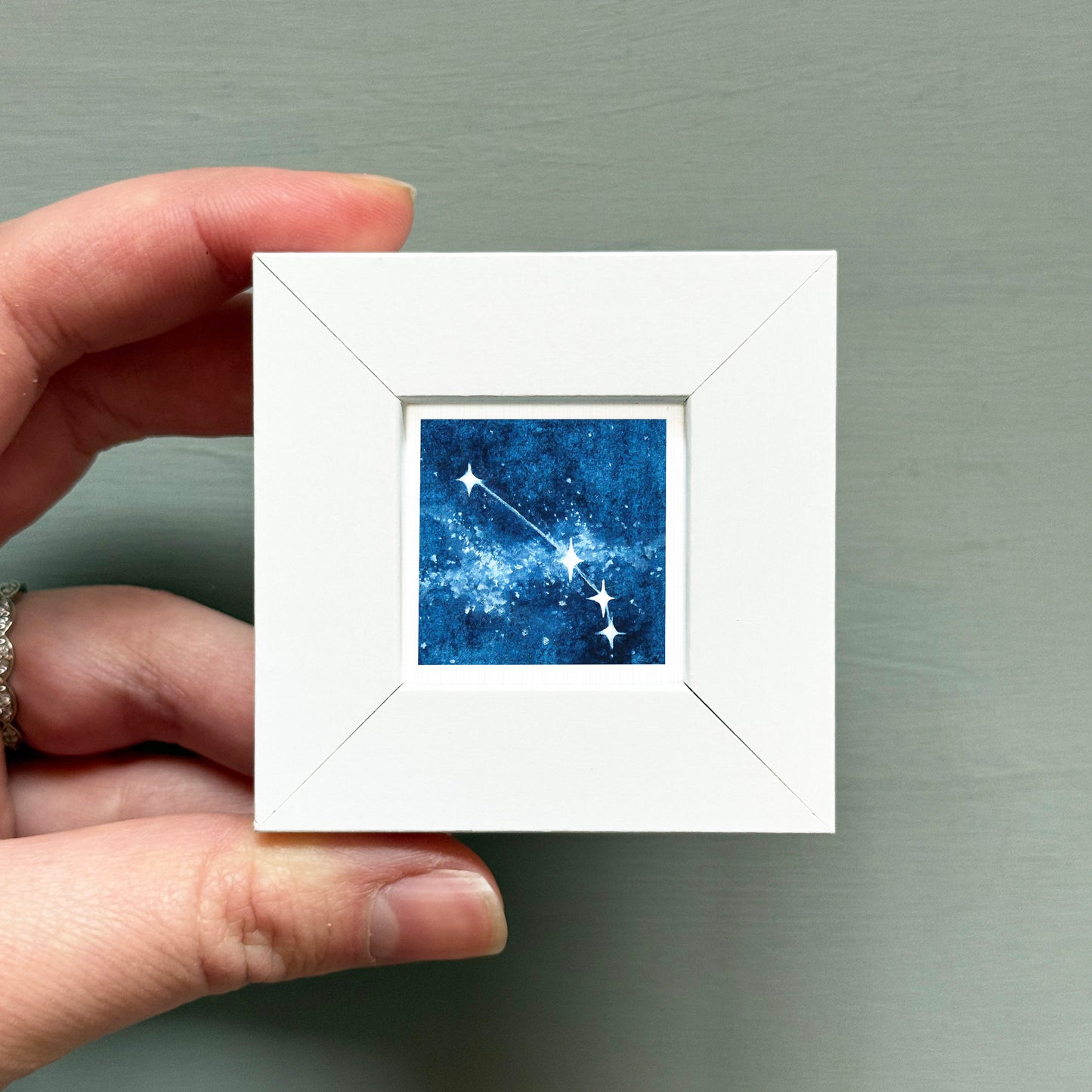 A hand is holding a small white frame containing a framed image of a starry night sky.