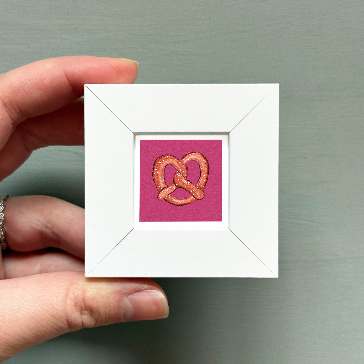 A hand is holding a small white square frame containing a framed image of a pretzel.