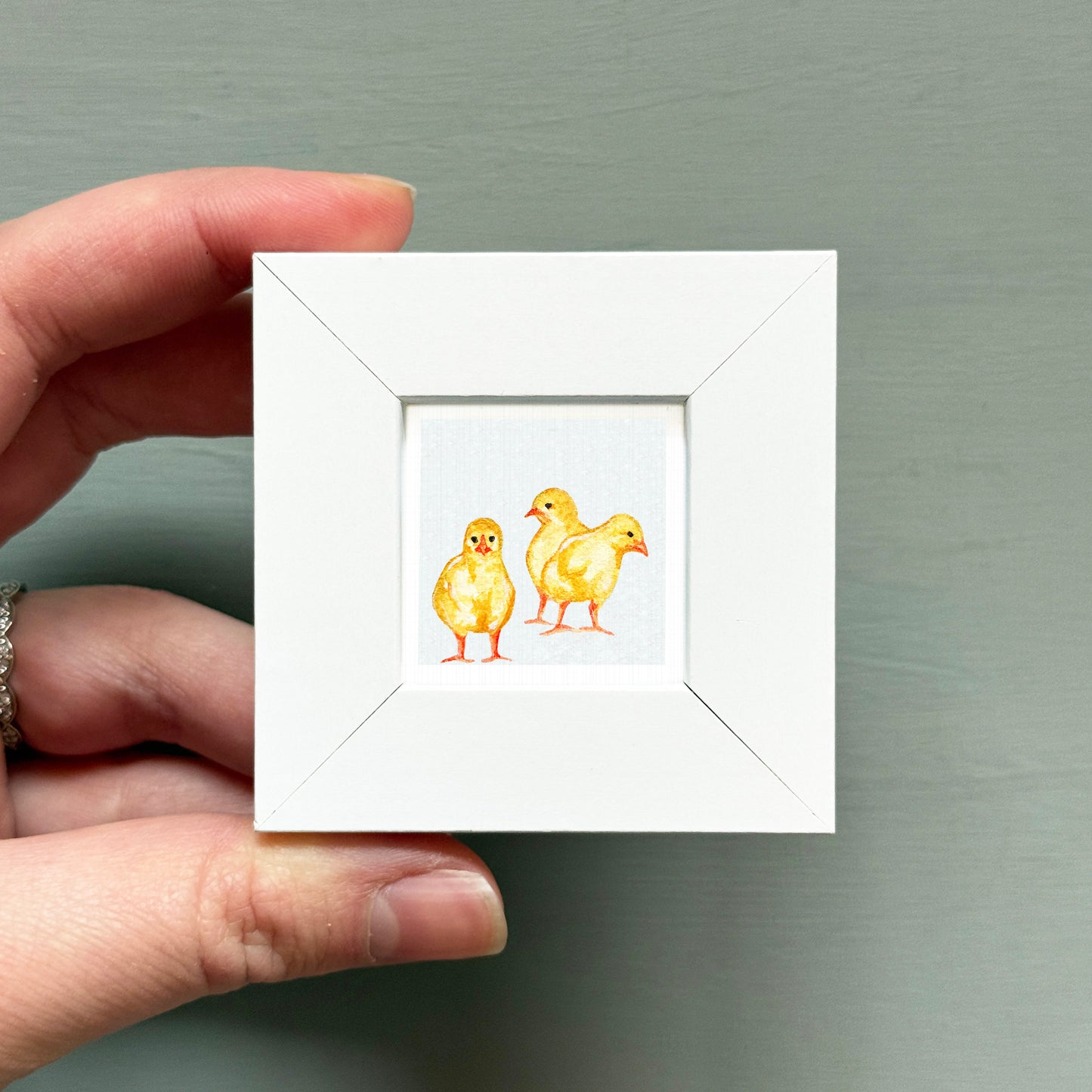 A hand is holding a small white square frame containing a painting of three yellow chicks.