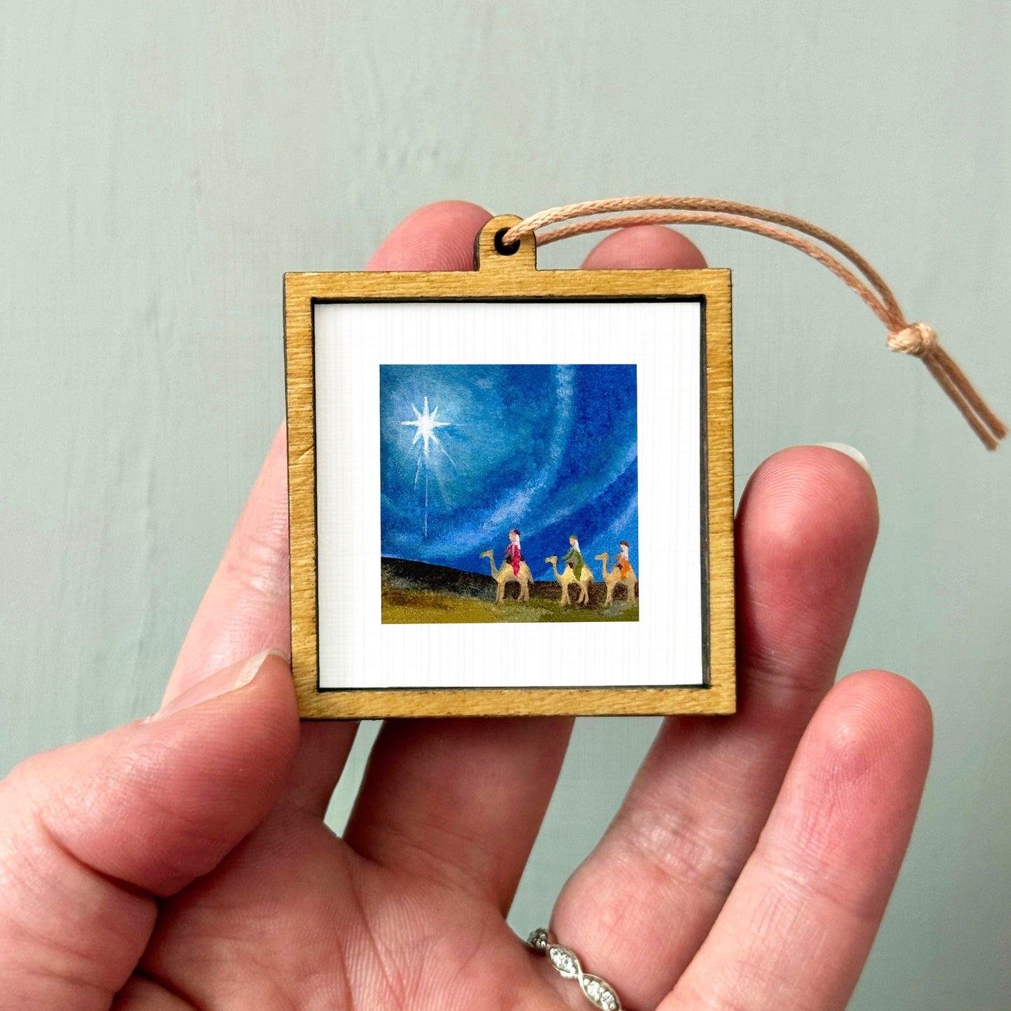 A hand holding a small wooden frame containing a painting of a starry night sky with a bright star and a group of people on a horseback.
