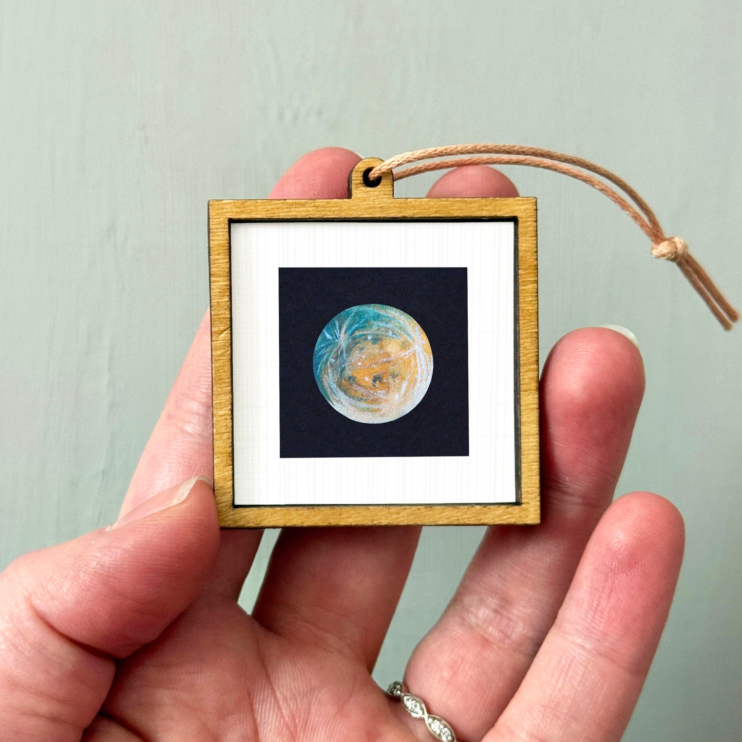A hand holding a small wooden frame containing a miniature image of a planet or celestial body.
