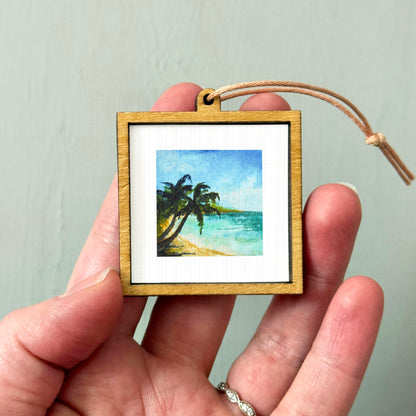 A hand holding a small wooden frame containing a painting of a tropical beach scene with palm trees.