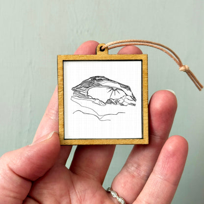 A hand holding a small wooden frame containing a black and white line drawing of a bird.