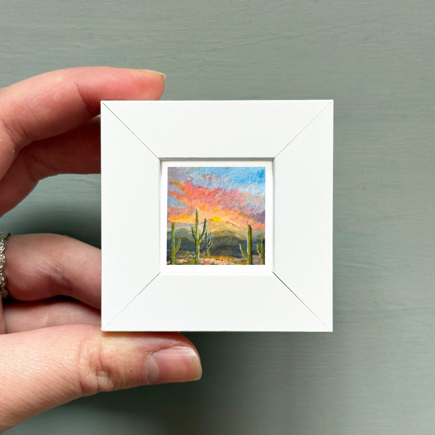 A hand is holding a small white square frame containing a painting of a desert landscape with cacti and a colorful sunset sky.
