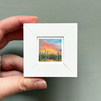 A hand is holding a small white square frame containing a painting of a desert landscape with cacti and a colorful sunset sky.