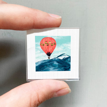 A person's hand is holding a small square picture depicting a hot air balloon floating above a mountain landscape.