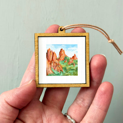 A hand holding a small wooden frame containing a watercolor painting of a mountain landscape.