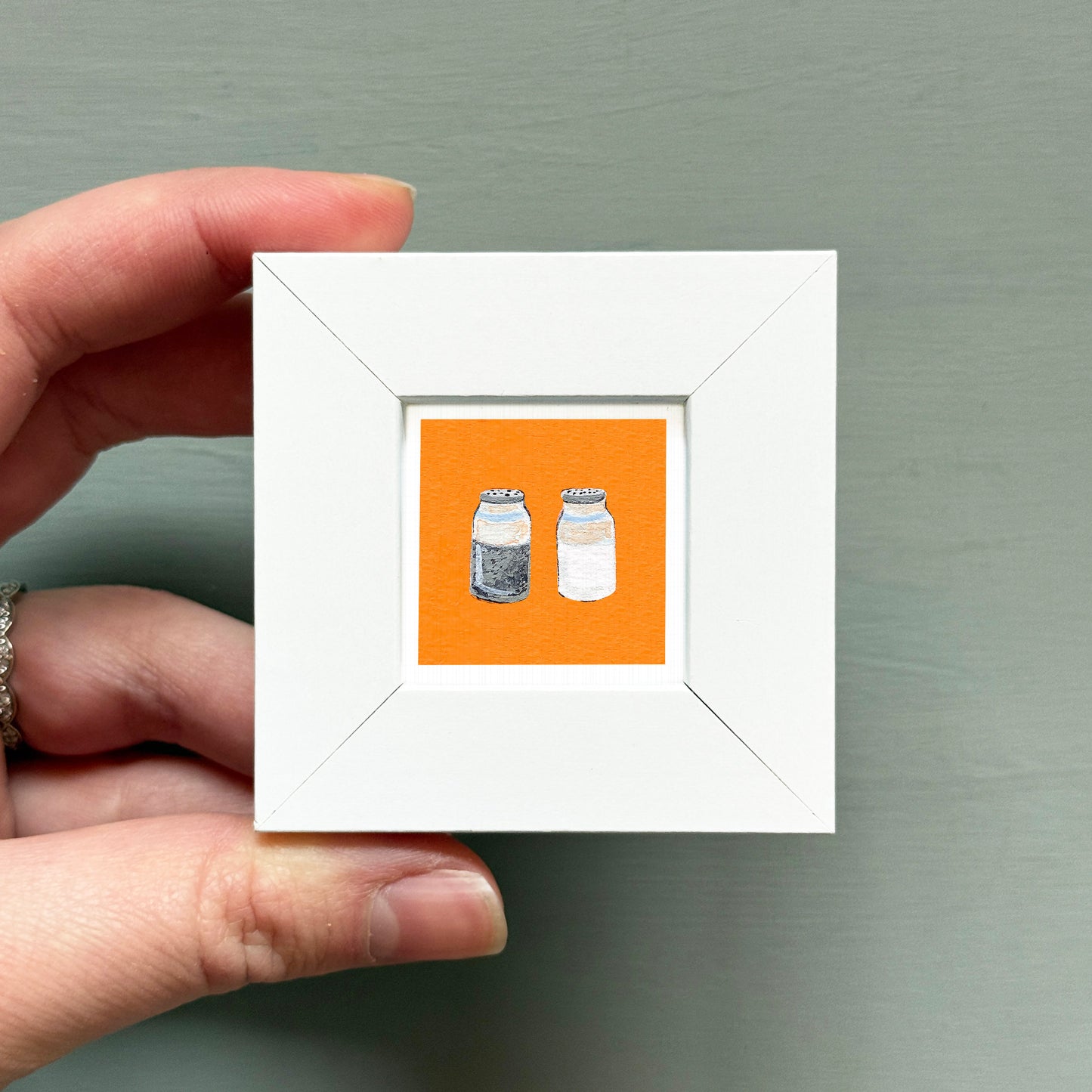 A hand is holding a small white square frame containing an illustration of two identical jars against an orange background.