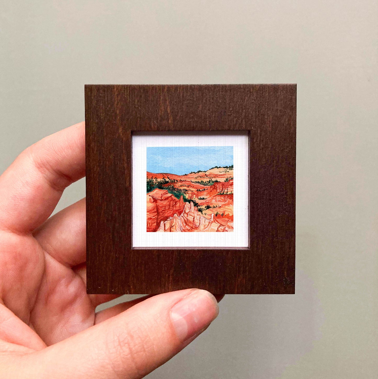 A hand is holding a small, square-shaped wooden frame containing a miniature painting of a desert landscape with red rock formations and a blue sky.