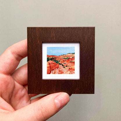 A hand is holding a small, square-shaped wooden frame containing a miniature painting of a desert landscape with red rock formations and a blue sky.