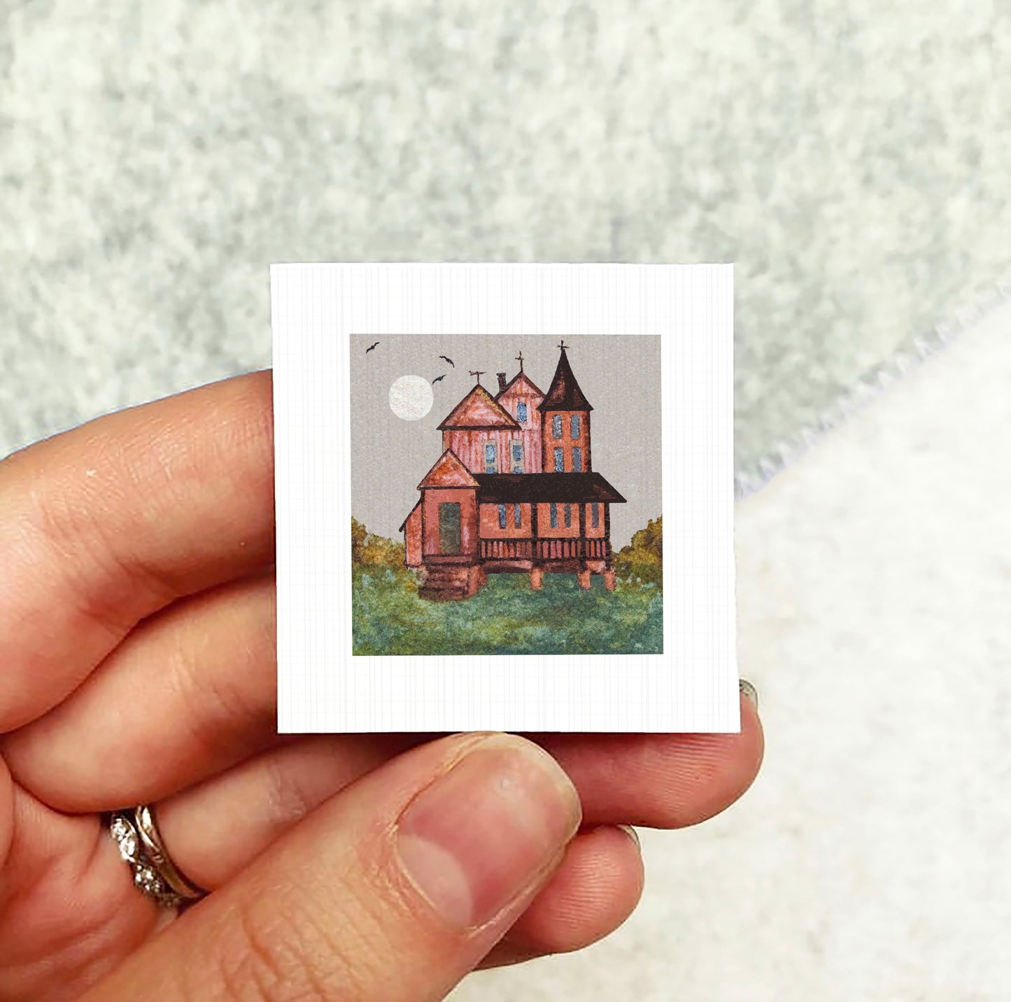 A hand holding a small square picture depicting a house with a red roof and a white moon in the sky.