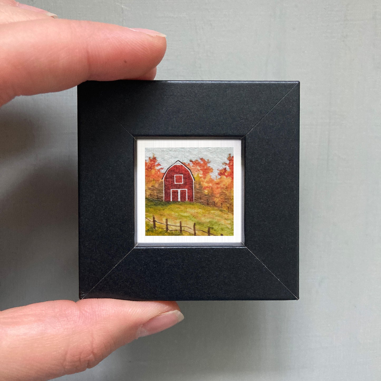 A person's hand holding a small black picture frame containing a miniature painting of a red barn surrounded by autumn foliage.