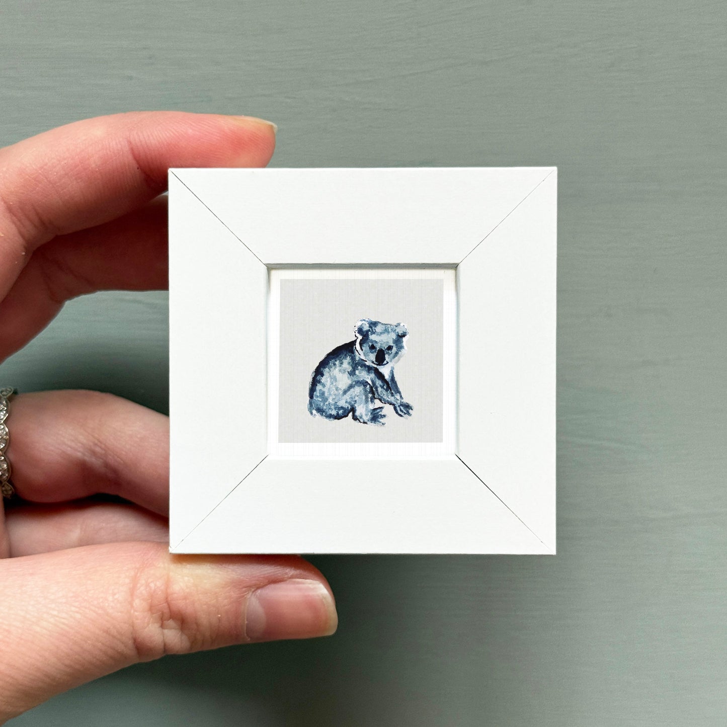 A hand is holding a small white square frame containing a painting of a koala bear.