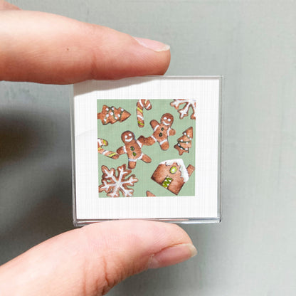 A person's hand holding a small square picture featuring various gingerbread cookies and snowflakes against a green background.