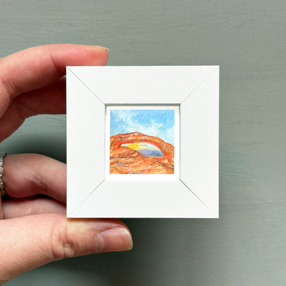 A hand is holding a small white square frame containing a watercolor painting of a desert landscape with a rainbow arching over it.