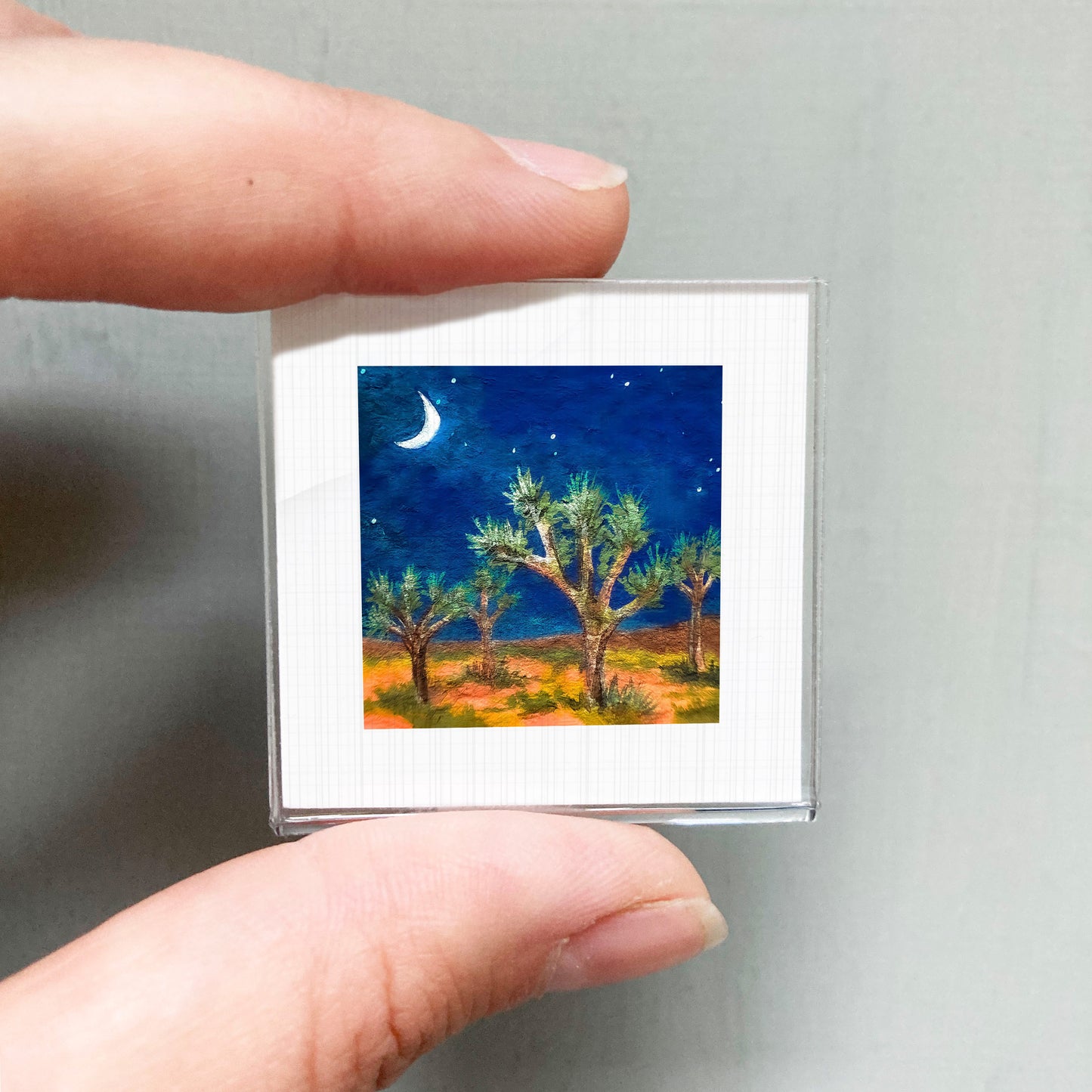 A person's hand holding a small square picture of a desert landscape at night, with a crescent moon and stars in the sky.