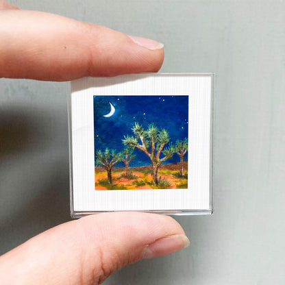 A person's hand holding a small square picture of a desert landscape at night, with a crescent moon and stars in the sky.