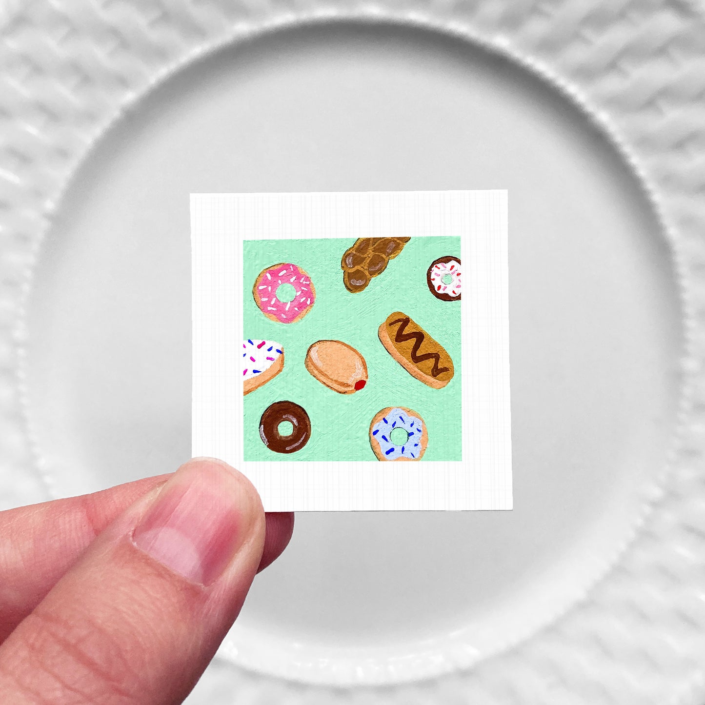 A hand is holding a small square card with a vibrant illustration of various donuts on it.
