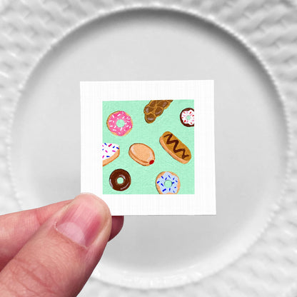 A hand is holding a small square card with a vibrant illustration of various donuts on it.
