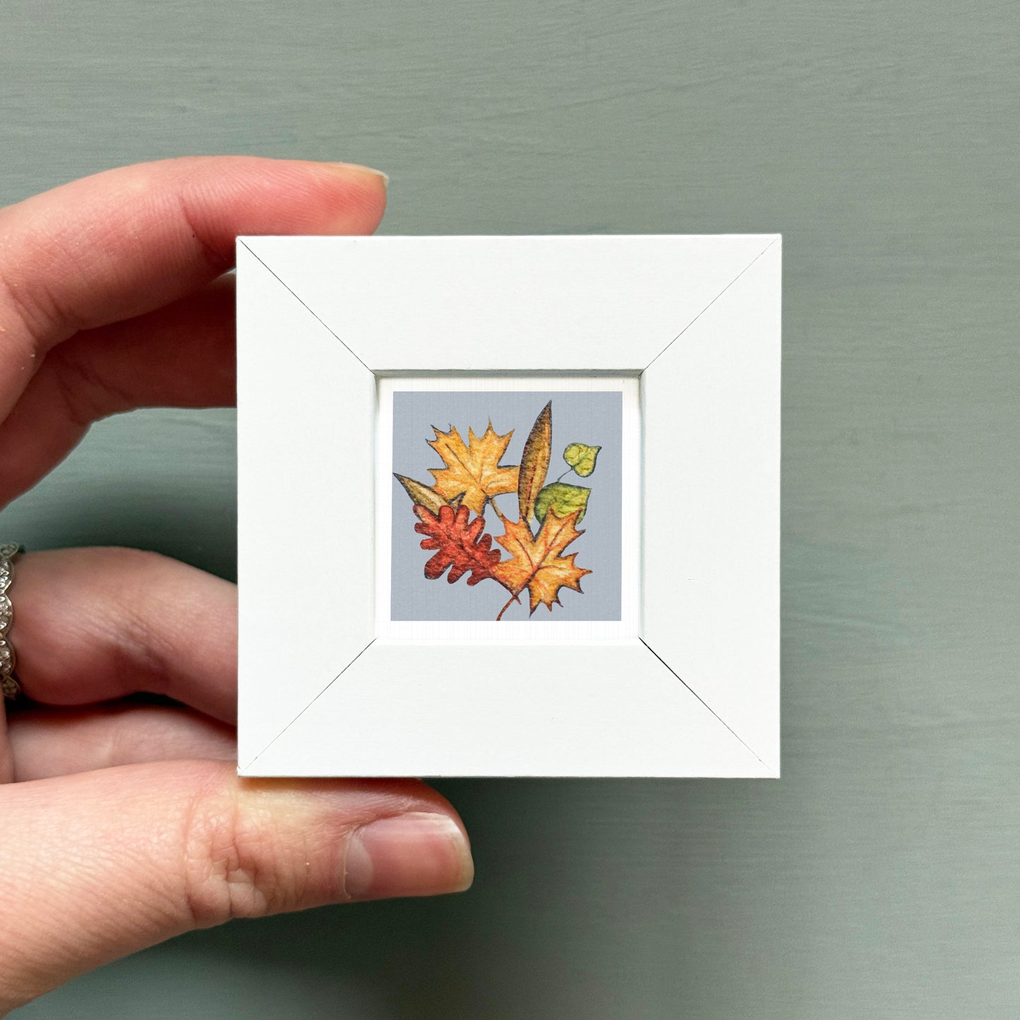 A hand is holding a small white square frame containing a colorful illustration of autumn leaves and a butterfly.