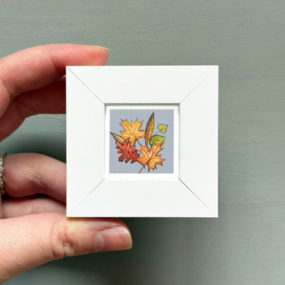 A hand is holding a small white square frame containing a colorful illustration of autumn leaves and a butterfly.