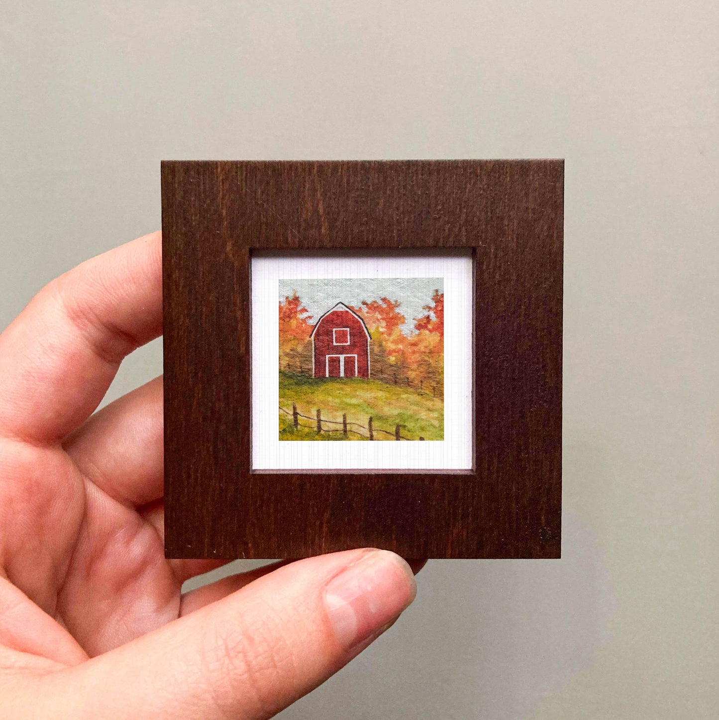 A hand is holding a small wooden frame containing a painting of a red barn surrounded by autumn foliage.