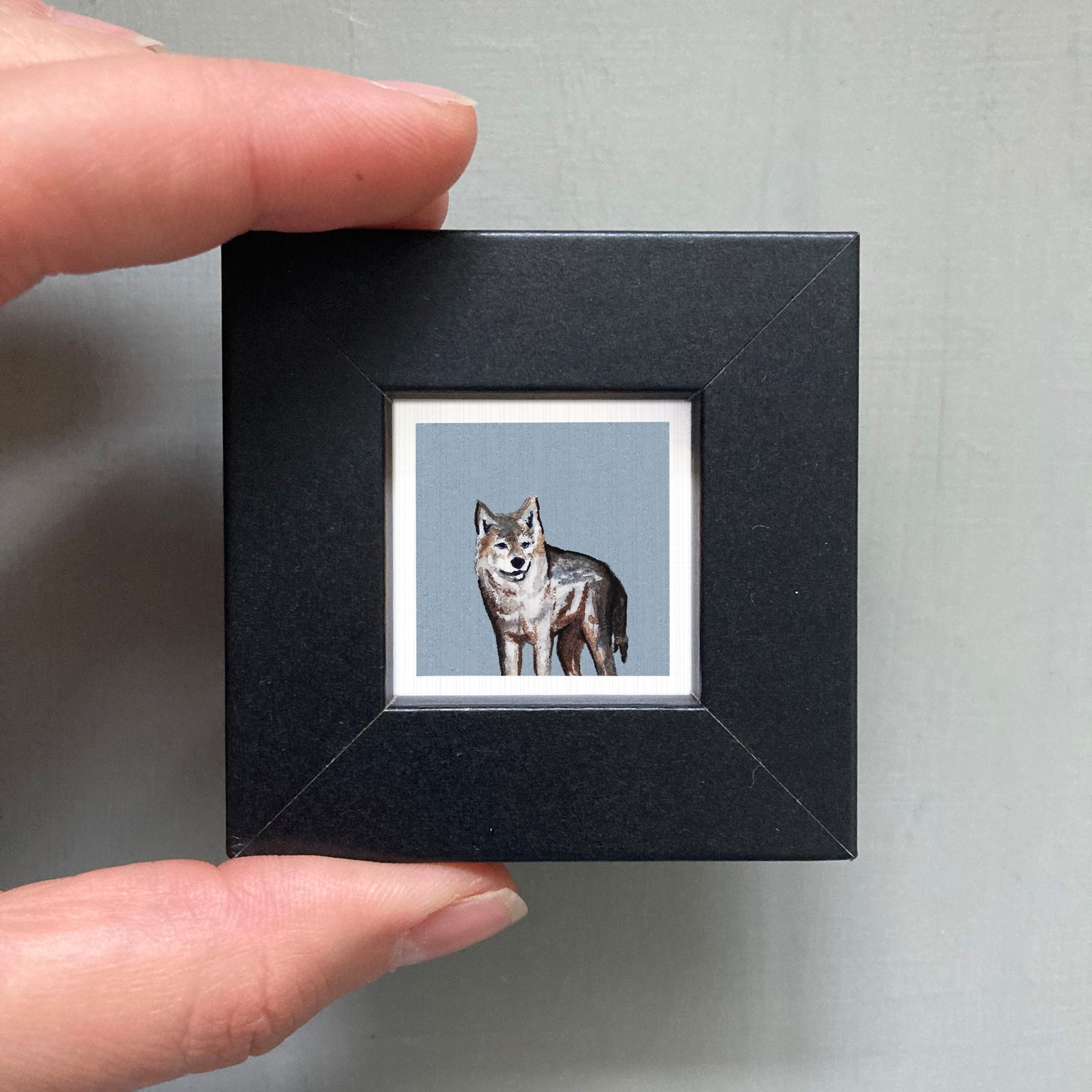 A person's hand is holding a small black frame containing a framed painting of a wolf.