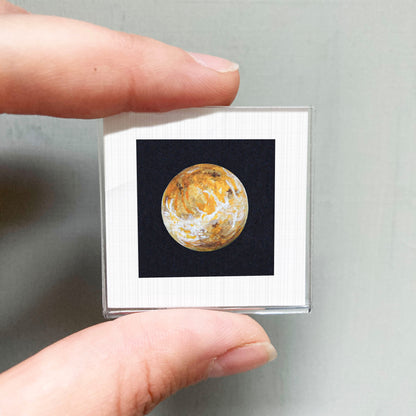 A person's hand is holding a small square frame containing a detailed image of a planet, likely Mars, against a dark background.