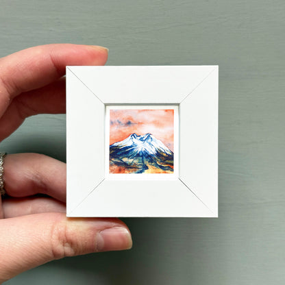 A hand is holding a small white picture frame containing a watercolor painting of a mountain landscape.