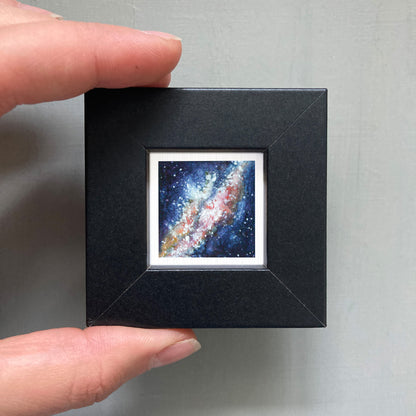 A person's hand holding a small, square-shaped black frame containing a miniature painting of a galaxy.