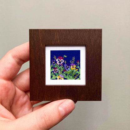 A hand is holding a small square picture frame containing a vibrant painting of colorful flowers against a dark background.