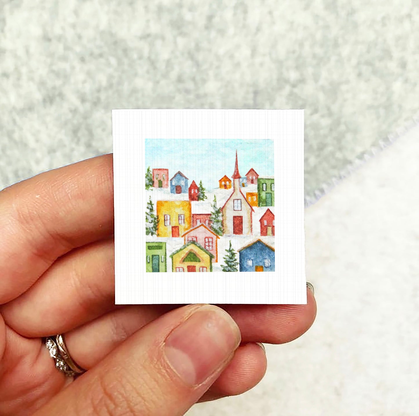A hand holding a small square-shaped paper with a colorful illustration of a winter village scene.